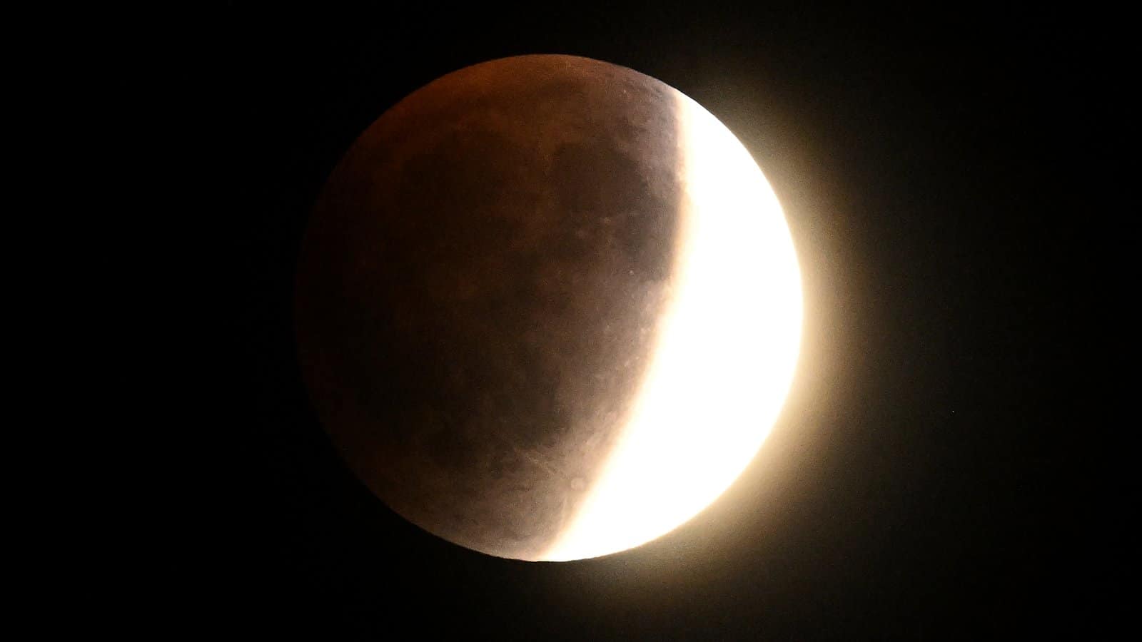 nature A full moon is chown with Earth's crescent shadow falling upon it during a total lunar eclipse. The extreme right edge is bathed in direct sunlight while the left is bathed in a rusty shadow.