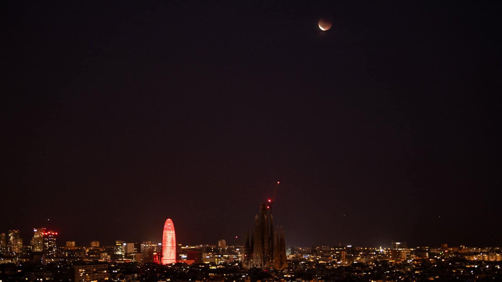 nature An almost fully eclipsed moon is pictured hanging in the dark sky over a city skyline. Tall buildings are visible near the center of the shot. The moon's lower crescent is brightly lit with the rest bathed in deeper shadow.