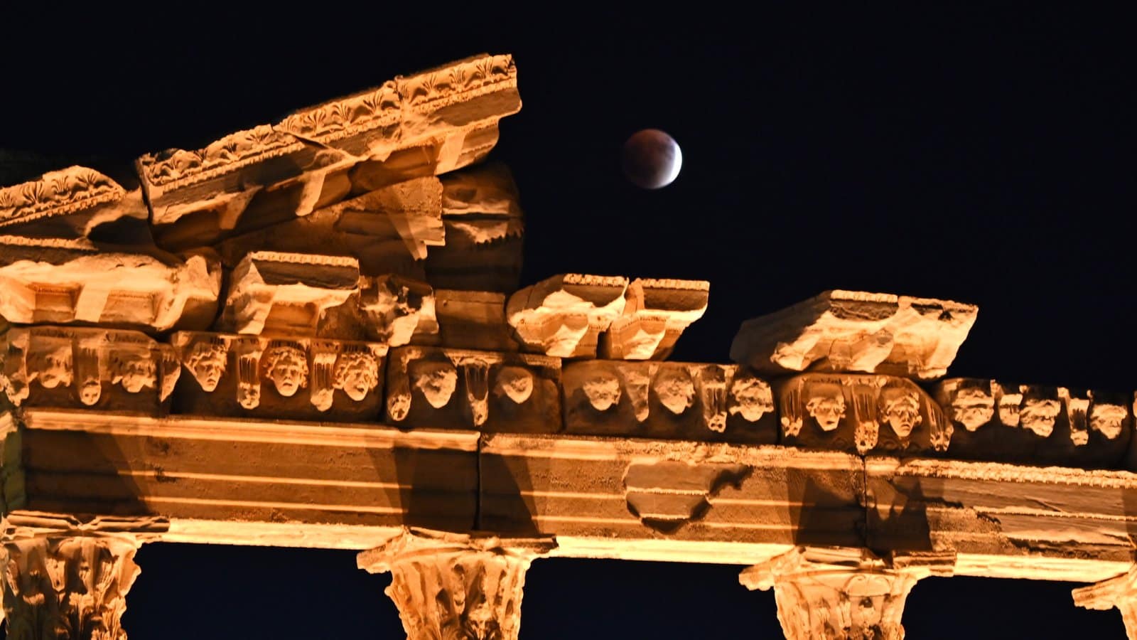 nature A mostly eclipsed full moon is pictured hanging above the collapsed roof of an ancient temple which is lit by yellow light. The moon's right crescent is lit, with the rest bathed in deeper shadow.
