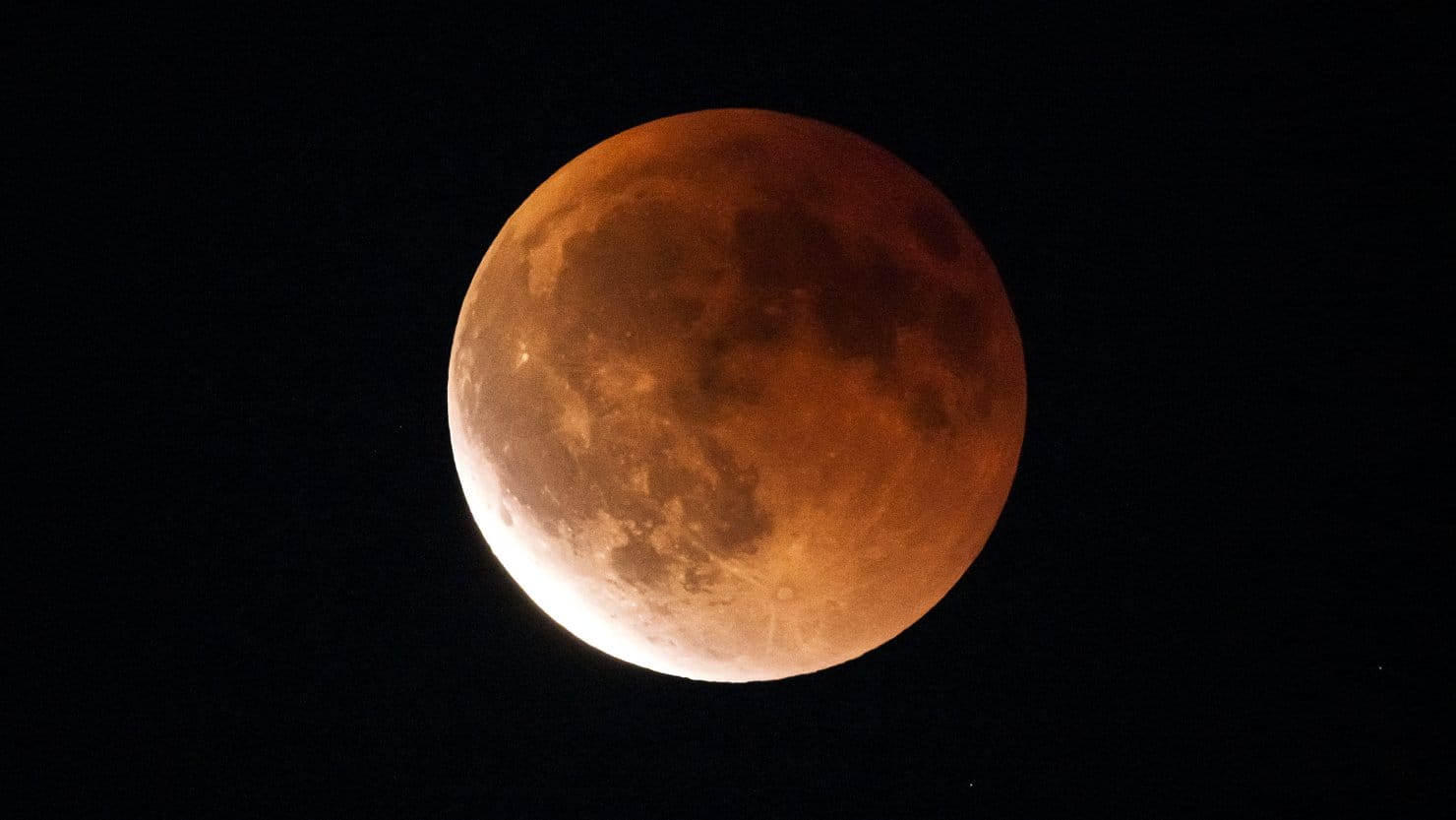 nature A yellow-orange eclipsed moon is pictured against a dark sky, with dark features known as lunar maria marking its surface along with the shapes of bright impact craters. The lower left part of its disk is brighter than the rest of its surface.